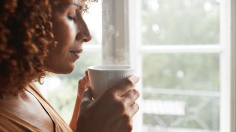 A woman closes her eyes as she holds a cup of coffee next to a window.