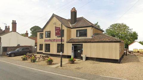 A two‑storey village pub clad in pale vertical timber, with a tiled roof and central chimney, facing a quiet road. A gravel forecourt contains wooden picnic tables and potted plants. Adjacent cottages and a small car park sit to one side, with trees and fencing behind.