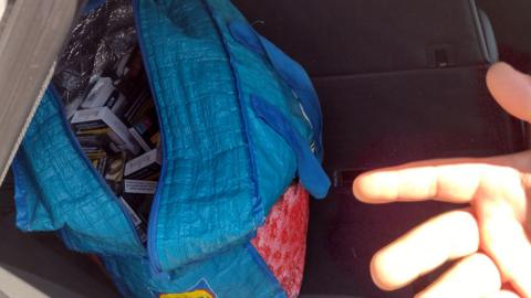 A blue bag full of cigarettes from the first seizure at the back seat of a car. A hand is showing pointing to it.