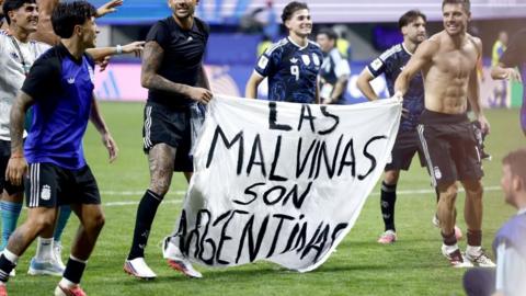 Argentina players hold a white banner that has "Las Malvinas son Argentinas", which translates as "The Falklands are Argentine", written in black text.