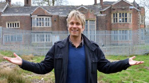 Hopwood Depree posing in front of Hopwood Hall, a large historic red-brick stately home, which is surrounded by a tall metal security fence