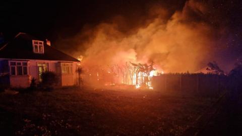 A small wooded area and a garden fence are seen on fire with large plumes of smoke coming away from the area. A house to the left of the blaze also appears to be on fire but only slightly. The photo has been taken at night. 