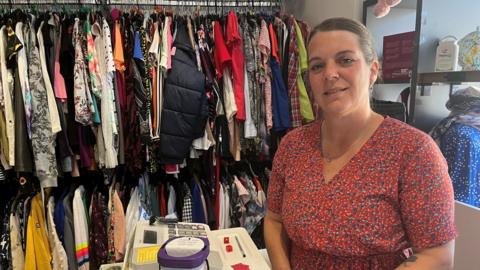 Emma stood in one of her community shops. She is wearing a red flowery dress and is stood in front of clothes selling the shop.