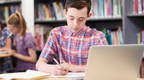 Male GCSE student working at laptop in school library revising with past papers