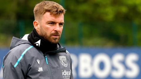 James Morrison wearing a grey West Bromwich Albion tracksuit top and black snood while in a training session with the word "BOSS" seen on a blurred advertising board in the distance behind him.