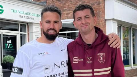 Mike is stood with his left arm around the shoulder of his friend Craig Eskrett. Mike is wearing a white T-shirt with company names and logos on acting as sponsors. He has a black goatee beard and swept-back hair. Craig has a burgundy Hull KR hoodie and has short dark hair. The pair are stood in front of a row of shops and looking into the camera.