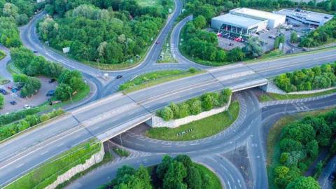 Running Horse Roundabout, near Maidstone. Grey roads leading onto a roundabout surrounded by green trees.