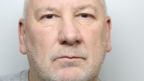A custody picture of a man with short grey hair and a beard. He is wearing a grey T-shirt.