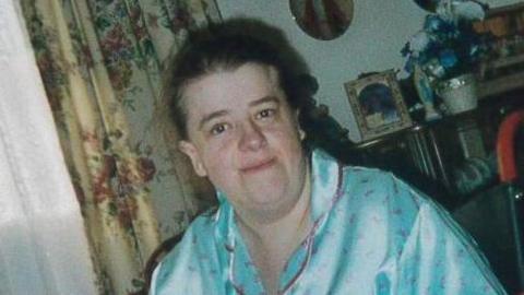 Angela Lloyd smiles at the camera. She is sitting in her wheelchair and is wearing blue silk pyjamas and appears to be in her living room with floral curtains and various knick-knacks on shelves behind her.