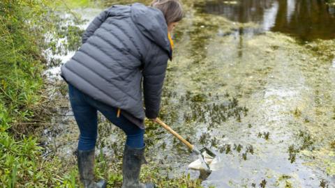 Jilly stands at the edge of the pond wearing black welly boots over jeans. She holds 