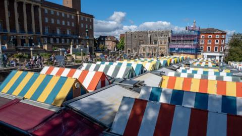 A view from above of Norwich market. We can see several brightly coloured, striped roofs. In the background prominent Norwich buildings can be seen, including City Hall.