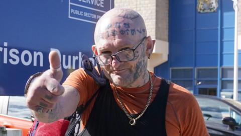Hoganson gives a thumbs up to the camera as he leaves HMP Durham. He is bald with symbols tattooed all over his head, neck, arm and hand, and is wearing an orange T-shirt with a black vest over the top. Behind him is a sign saying HMP Durham and a car park large blue wall.