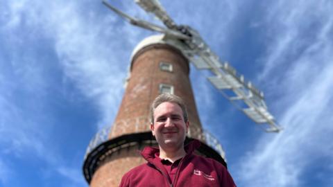 A man in a maroon uniform of polo shirt and fleece with English Heritage logos smiles down at the camera. Behind him stands the looming tower of a six sailed widnmill. It is built in red brick and has a wooden platform halfway up and white sails.