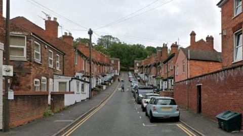 View of Vine Street in Lincoln. It is a row of red brick terrace houses along a hill with trees at the end. Cars are parked alongside the right-hand side of the road. 