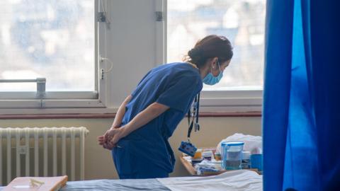 A nurse wearing blue scrubs looks down at a wooden table next to a patient's bed. The bed is empty and has been made up. A blue curtain hangs next to the bed.