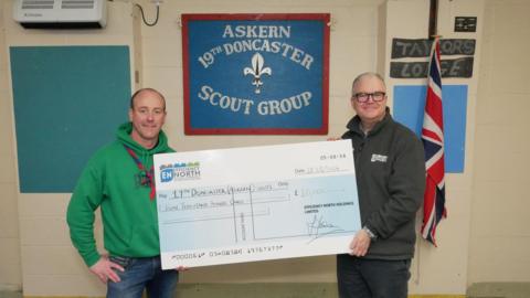 A man wearing a green hoodie and a blue and red knecker and a man in a grey fleece with glasses hold a giant cheque for £10,000.