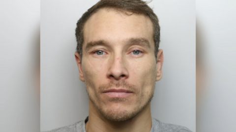 A close-up police mug shot of Lewis Moore on a white background. He has brown hair and a stubbly beard, and is wearing a grey T-shirt. 