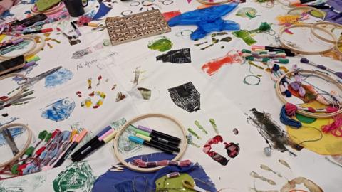 A white tablecloth covered with arts and craft supplies - pens, paints, wool, letter blocks - and artwork. 