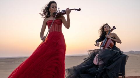 Two women outdoors in the desert, playing the violin and the cello. One woman is standing up, wearing a red dress, the other is sitting down and wearing a black dress. 