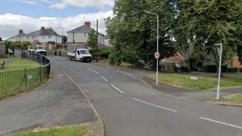 The entrance to a road of houses, with a park to the left and patch of grass to the right