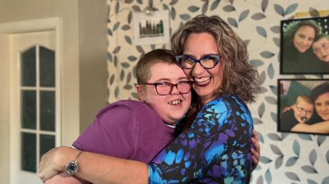 A woman and a boy are hugging. The woman is wearing a blue and purple patterned blouse and the boy is wearing a pink t-shirt. On the wall behind them, there are photos of the woman and the boy when they were younger.