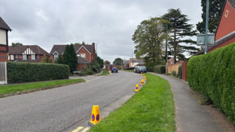 A road has yellow traffic cones along the right hand side, next to a grass verge and a pavement. There are houses either side of the road and cars parked and driving in the distance.