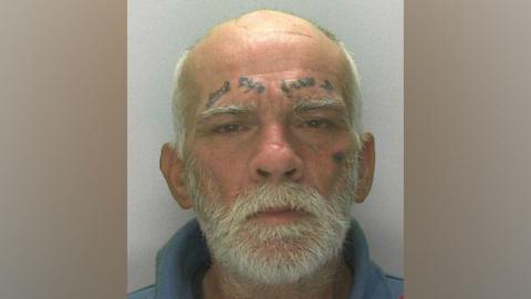 A custody picture of Anthony Gardiner. He is wearing a pale blue fleece or shirt with the collar high around his neck, and has a white beard, thick white eyebrows and balding white hair. He is looking at the camera and has faded tattoos above both eyebrows and one small one beneath his left eye.