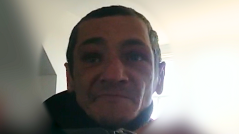 Ciprian Ilie as seen via a police bodyworn camera. He has short dark hair and stubble. He is looking slightly resigned as he gets arrested. The background is blurred.
