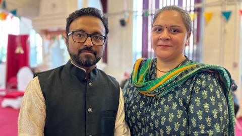 Ashish and Meera standing in a room in the temple, looking directly at the camera. Ashish has short black hair and a neatly trimmed beard and moustache. He has glasses and is wearing a white kurta and a black waistcoat. Meera has tied back grey hair and is wearing a blue and yellow Indian dress with a stole around her neck.