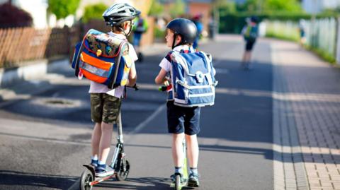 Two young boys on scooters wearing backpacks leaving school in hot sunny weather.