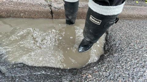 A person with wellington boots stands inside a pothole at the side of a Derbyshire road in March. The water is grey and the surrounding road black and the water level covers the top of the foot part of the boots.