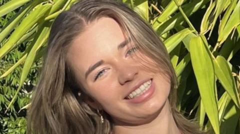 Holly Bowles smiles in front of a green plant