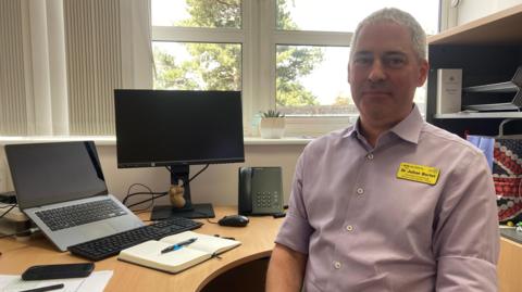 A man with short grey hair sits in an office, with a curved desk under a window and a shelving unit behind him. He is wearing a light purlple shirt with a yellow name badge that reads Dr Julian Berlet.