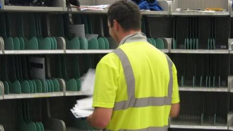 A postal worker wearing a yellow hi-vis with his back facing the camera. He is sorting post onto the shelves which have green dividers. 