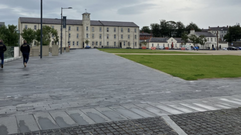 Shows Ebrington Square with pavement on the left and two people with their backs to the camera, grass on the right, a hotel and other buildings in the background.