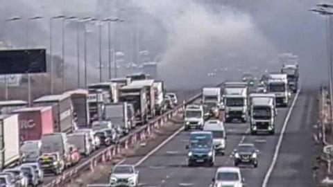 A motorway CCTV camera view shows traffic at a standstill on one carriageway of the M62. Grey plumes of smoke can be seen coming from a lorry, with the fire spreading to the side of the road. 
