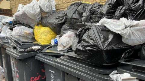 A row of six black bins up against the side of a brick house. They are overflowing with black, white and yellow bin bags. There are some bags on the wet floor, and others piled high on top of the bins. 