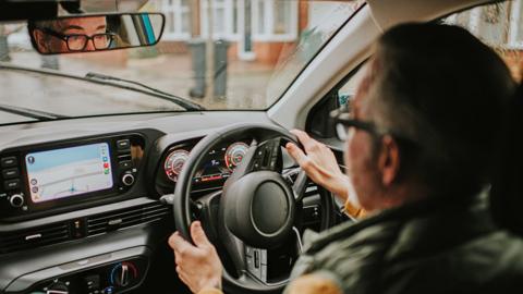 View over the shoulder of a male driver. He has is wearing glasses. It is a rainy day