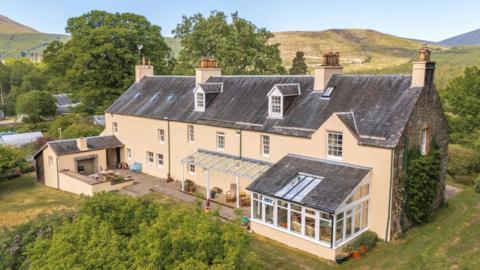 The house is of period design with a slate roof and cream-coloured walls. A gable end is covered in icy. It is set in a Highland landscape of rolling hills. 