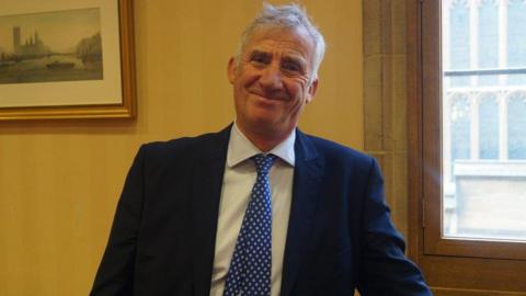 Lord Storey, who has grey hair and wears a blue suit over a white shirt, smiles at the camera 