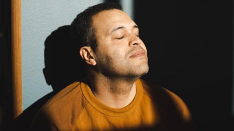 A man in an orange jumper with his head tilted back and his eyes closed with the light on his face.