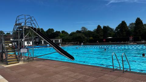 A bright blue outdoor swimming pool under a clear blue sky. There is a black slide into the pool and a ladder. There are people swimming in the pool, at the bottom end of which there is a white poolhouse. There are trees surrounding the site the lido is on.