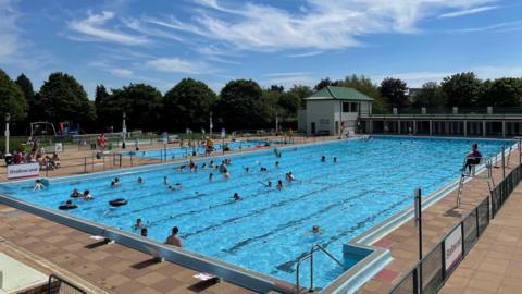 Outdoor swimming pool with a smaller pool in the background and a people swimming in them with a lifeguard sitting on the chair on the side of the pool