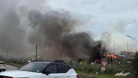 A view of a large smoke plume in the sky, flames at the bottom on large piles of rubbish. There are fences, a road and a couple of cars in the foreground.