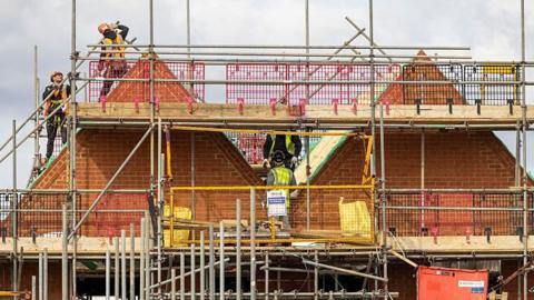 A partially constructed brick building surrounded by extensive metal scaffolding. Several construction workers wearing safety gear are working on the upper level near triangular roof structures. The site includes wooden planks, metal poles, and safety barriers in pink, grey and yellow colours. A cloudy sky forms the background.