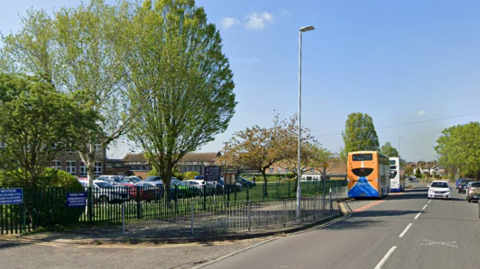 A road with two buses on one side and white car on the other. There is a lamppost, blue sky, green trees and a gated car park next to the road