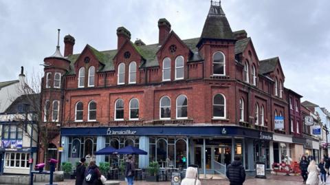 A three-storey red brick building with chimneys and turrets is at the side of an open, pedestrianised square in a town centre. A pedestrianised street runs along the side of the building on the right. There is a pub and hotel building next to it on the left. Shoppers are walking in front of it.