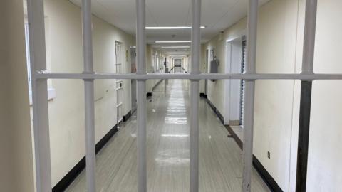 A barred door blocks the photographer's way. There is a grey floor and white walls lining a corridor heading straight into the back of the shot with a barred door to the left and white shutters to the right.