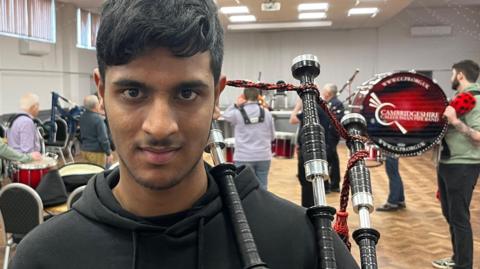 Yojit Bodduluri is 14 years old, has black hair and some whiskery stubble around his chin and under his nose, he is wearing a black fleece top and has his bagpipes on his left shoulder, behind him are other pipers and drummers, they are in a community hall, with chairs and tables around too.
