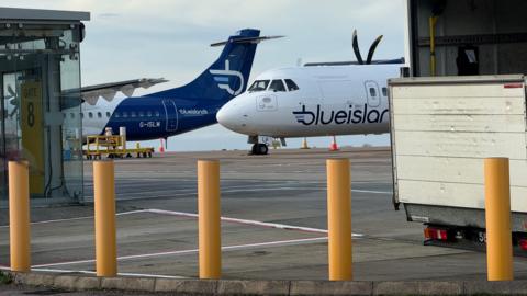 Two Blue Island planes on the tarmac at an airport. Large yellow bollards are in the foreground of the photo. The back of a lorry parked on the runway can be seen just behind the bollards.